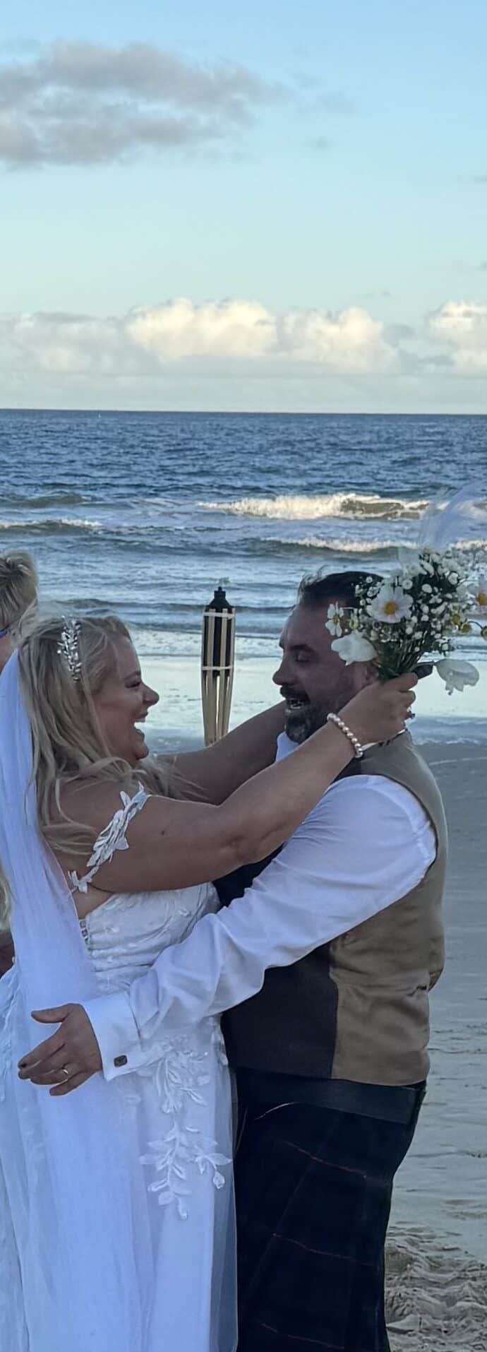 A bride and groom embrace joyfully on a sandy beach, with the bride holding a bouquet above the groom’s head. The ocean and a cloudy sky are in the background, and another woman smiles nearby.