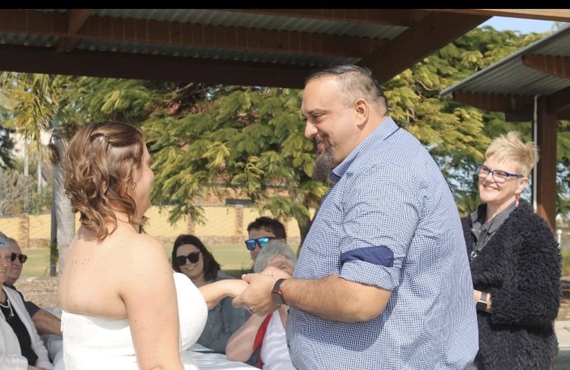 A bride and groom hold hands and smile at each other during an outdoor wedding ceremony, while guests and an officiant look on under a pavilion. Trees and sunlight can be seen in the background.